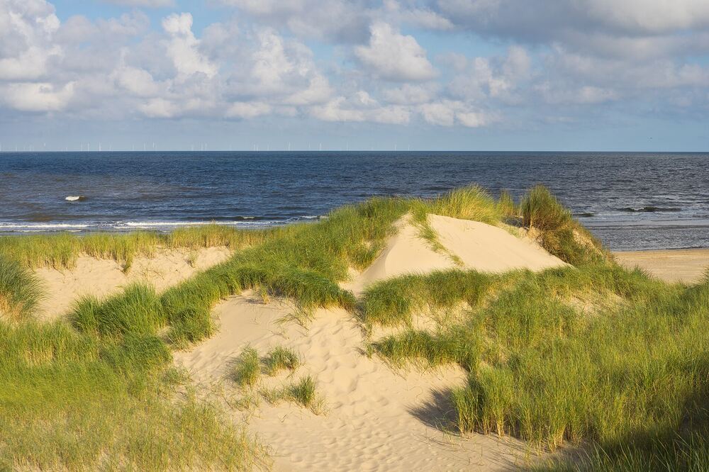 Zandduin met helmgras en daarachter de blauwe Noordzee onder een lucht met wolken